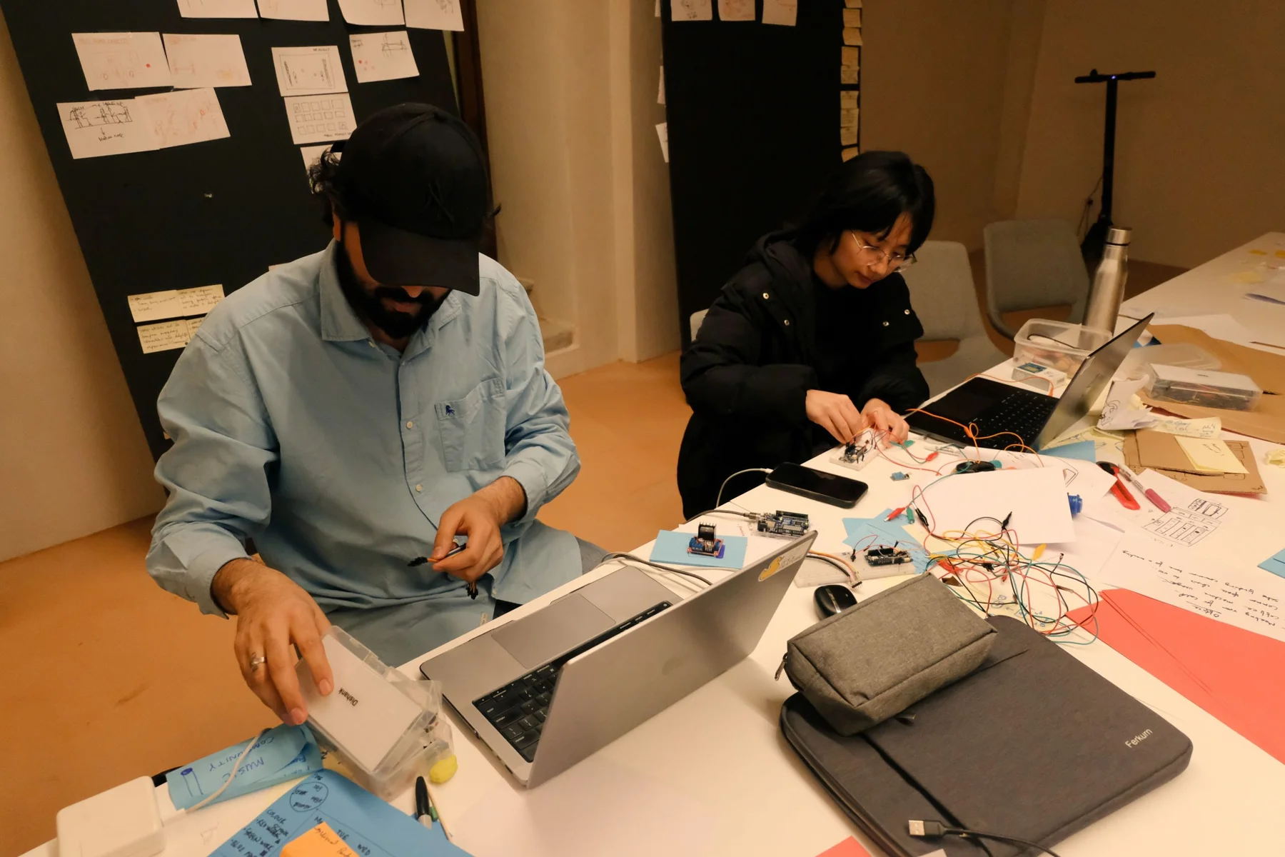 Two people building circuits at a table covered with wires, sketches and laptops