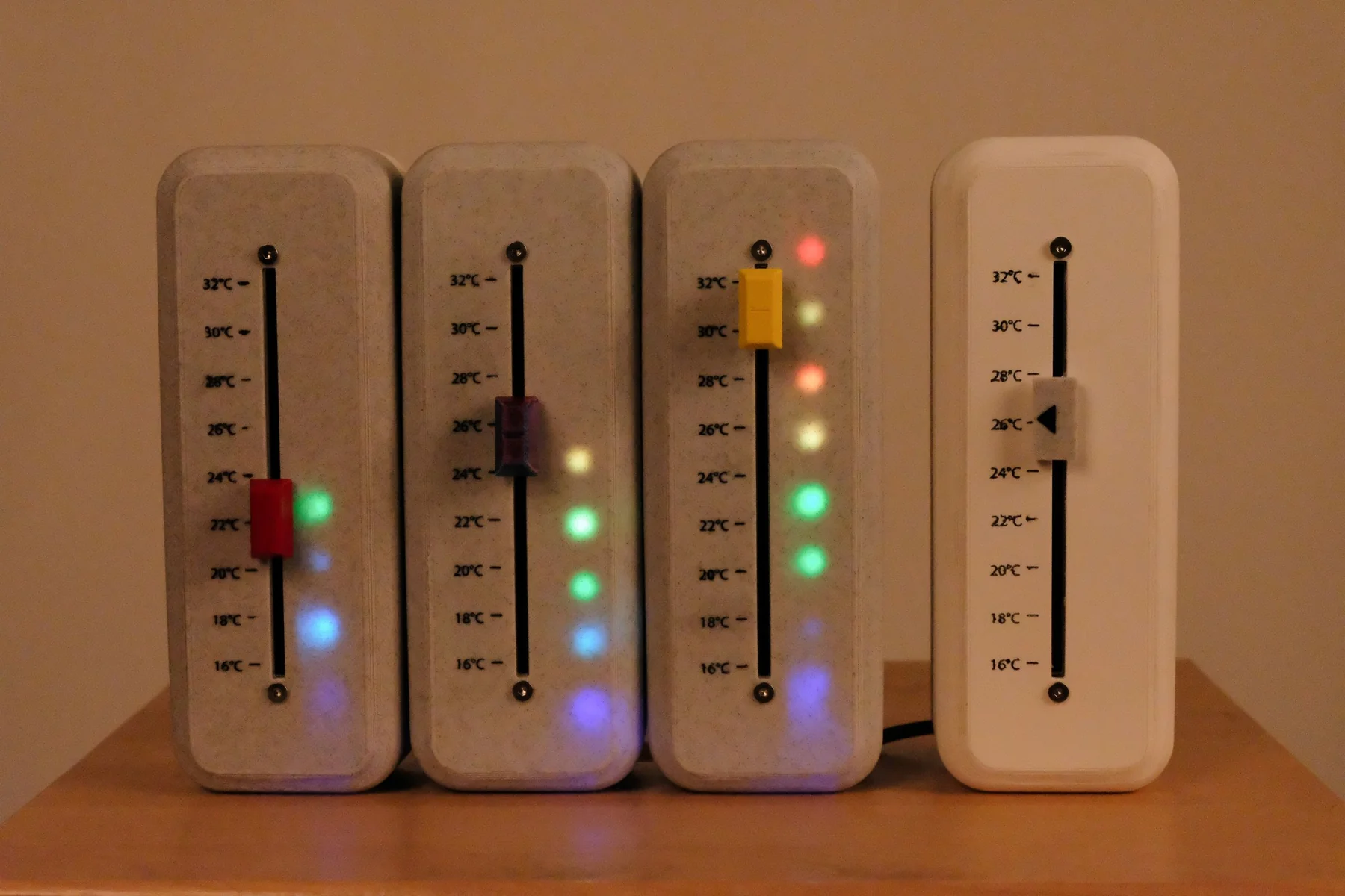Four thermostats side by side: three lit with coloured LEDs showing each fader position, and a plain white one